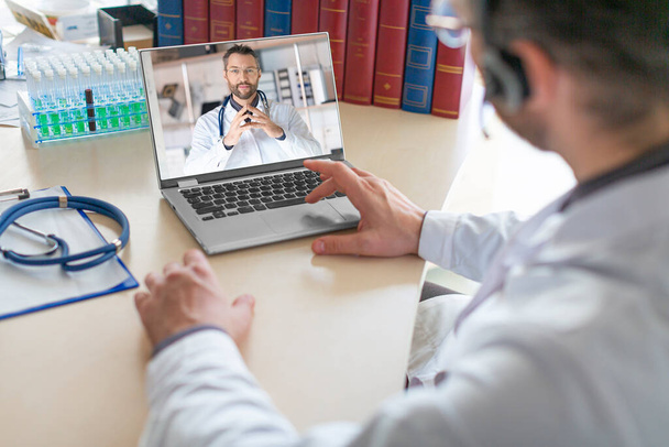 Un medico si siede nel loro studio medico e conduce una consultazione virtuale con un collega utilizzando un computer portatile. I medici stanno discutendo una storia clinica dei pazienti e le opzioni di trattamento. - Foto, immagini
