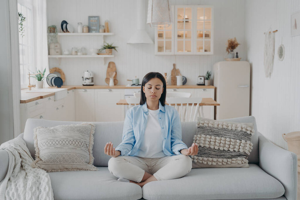 Peaceful mind, zen and concentration. Young hispanic woman is practicing yoga at home. Girl sitting on couch in lotus pose and meditating with her eyes closed. Emotion balance and stress relief. - Valokuva, kuva