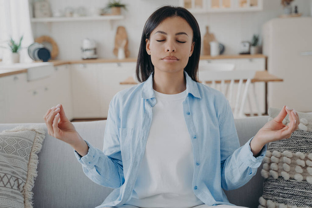 Спокойная женщина медитирует сложенными пальцами на жесте мудра, сидя дома на диване. Мирная молодая женщина практикует йогу, снимает негативные эмоции, расслабляется на диване в гостиной. Управление эмоциями. - Фото, изображение