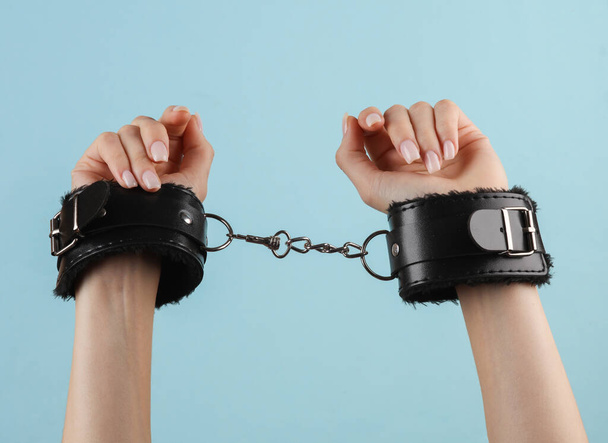 Women's hands in leather handcuffs on blue background. BDSM accessories, domination, sex games - Photo, Image