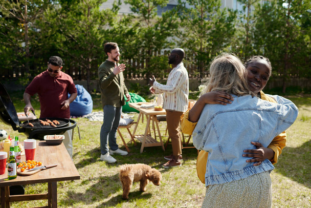 Счастливые молодые межкультурные женщины обнимают друг друга против двух мужчин, обсуждающих и их друга, делающего барбекю на гриле - Фото, изображение