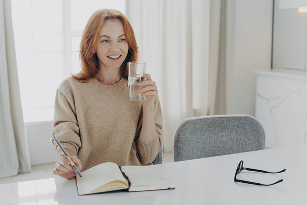 Happy smiling red haired woman housewife dressed casually with glass of pure mineral water enjoying morning at home, writing shopping list in notebook and planning her day, looking aside with smile - Fotoğraf, Görsel