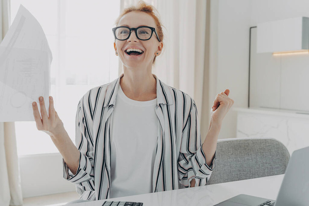 Excited young woman with paper bills in hands feeling euphoric, happy about finally paying down her mortgage debt, exclaiming yes with excitement while sitting at table with laptop - Photo, Image