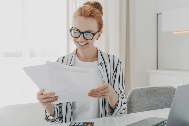 Happy smiling caucasian woman reading good news in financial documents while managing family budget, ginger female receiving bank notification while sitting at table in kitchen with laptop, calculator - Photo, Image