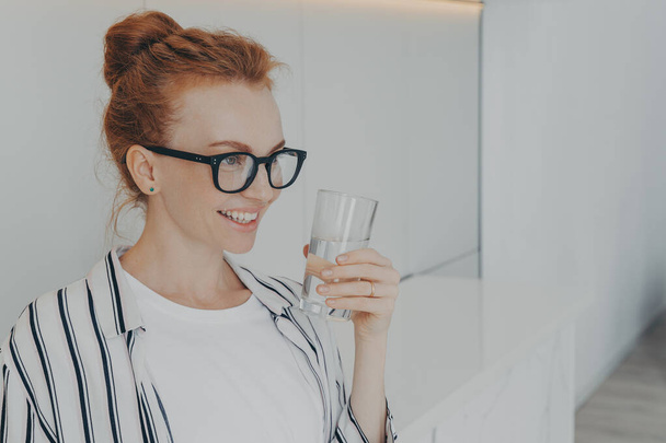Indoor shot of smiling redhead young woman holds glass of water drinks aqua for body refreshment follows healthy lifestyle wears spectacles striped shirt focused into distance. Hydration concept - Photo, Image