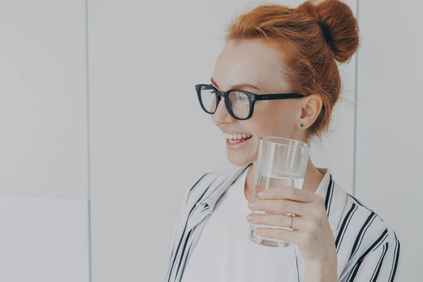 Thirsty woman with red hair combed in bun drinks still water prevents dehydration holds glass maintains balance of body leads healthy lifestyle wears spectacles striped shirt looks away pensively - Valokuva, kuva