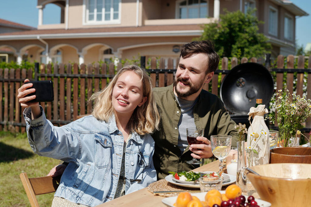 Νεαρό χαρούμενο ζευγάρι ή φίλοι που κάνουν selfie στο smartphone κατά τη διάρκεια υπαίθριου δείπνου ή πάρτι ενώ κάθονται δίπλα στο σερβιρισμένο τραπέζι - Φωτογραφία, εικόνα