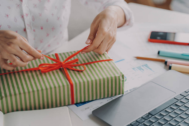 Cropped image of young woman office worker decorates Christmas gift with ribbon for friends or family members while sitting at desk in front of laptop at her workplace. New Year and winter holidays - Foto, Imagem