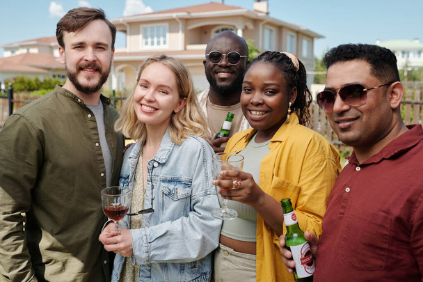Fröhliche junge interkulturelle Freunde mit alkoholischen Getränken blicken in die Kamera, während sie sich im Freien vor einem Landhaus treffen - Foto, Bild