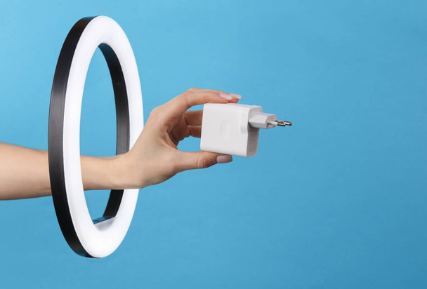 Woman's hand holds charger adapter through led ring lamp on blue background. Creative idea. - Photo, Image