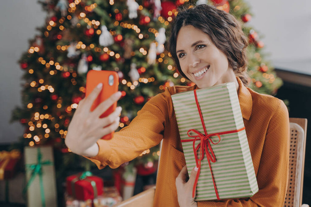 Újévi ünnepség. Vidám, vidám olasz nő ül közel gyönyörű díszített karácsonyfa otthon, kezében csomagolt karácsonyi ajándék doboz és pózol szelfi, boldog nő élvezi a téli ünnepek - Fotó, kép