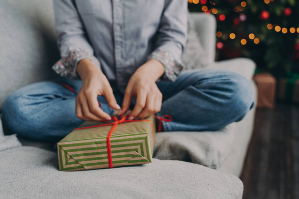 Foto cortada de mulher abrindo caixa de presente de Natal em casa, sentado no sofá perto da árvore de xmas decorada, presente de ano novo envoltório feminino durante as férias de inverno - Foto, Imagem