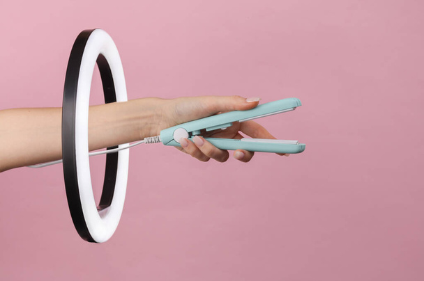 Γυναικείο χέρι κρατά ίσιωμα μαλλιών μέσω LED λαμπτήρα δαχτυλίδι σε ροζ φόντο. Δημιουργική ιδέα. Έννοια ομορφιάς - Φωτογραφία, εικόνα