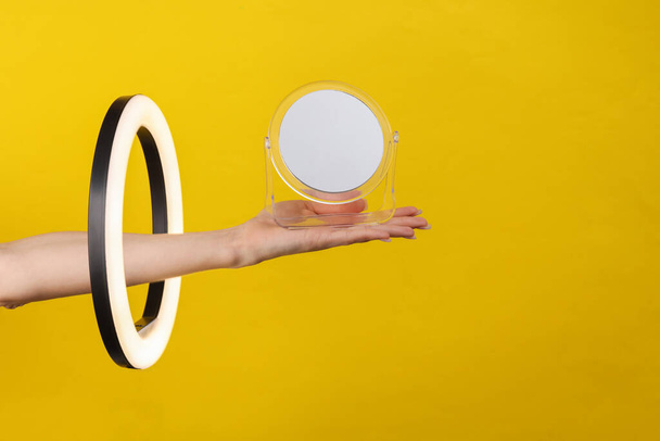 La mano de la mujer sostiene el espejo a través de la lámpara de anillo led sobre fondo amarillo. Una idea creativa. Concepto de belleza - Foto, imagen