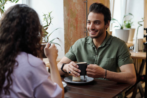Young smiling man and his girlfriend with cups of coffee sitting by table in front of one another and chatting while having cappuccino - Photo, Image