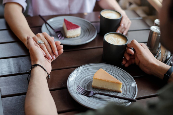 Руки молодых ласковых свиданий сидят за столом друг напротив друга и пьют кофе с вкусными ванильными и черничными чизкейками - Фото, изображение