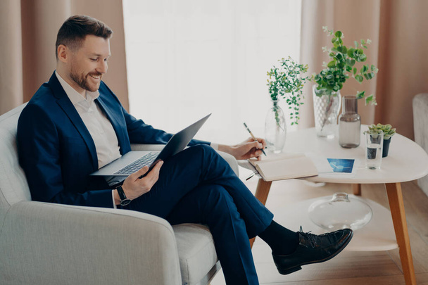 Boldog szakállas vezérigazgató online találkozót tart, miközben karosszékben ül a nappaliban, laptopozik. Sikeres üzletember öltönyben figyeli az üzleti webináriumot és jegyzetel, miközben beltérben pihen. - Fotó, kép