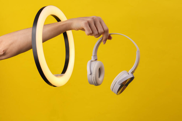 Die Hand der Frau hält weiße Kopfhörer durch eine LED-Ringlampe auf gelbem Hintergrund. Kreative Idee. - Foto, Bild