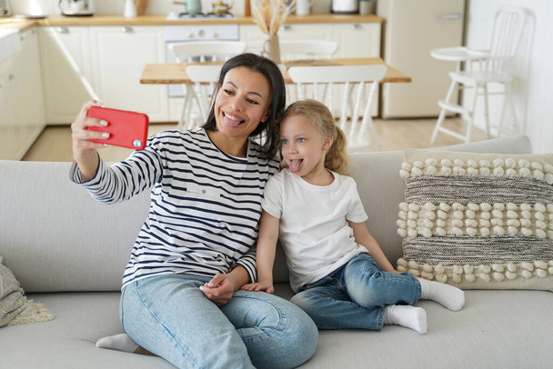 Mom and child are taking selfie on phone. People are having fun and showing tongues in front of camera. Young caucasian mother makes photo with little daughter on couch. Cheerful mum and kid at home. - Фото, изображение