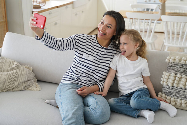 Funny little daughter and young mother showing tongue taking pictures of family, looking at mobile phone camera, happy mom having fun with kid girl, taking selfie together, sitting on couch at home. - Foto, Imagem