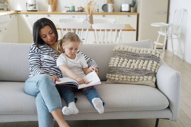 Мать с маленькой дочкой читает вместе книгу, учится, наслаждается семейным хобби. Мама обнимается, воспитывает девочку, сидя на диване, читает сказку дома. Родительство, материнство. - Фото, изображение