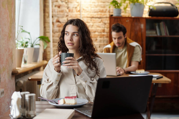 Mladý klidný žena na volné noze v casualwear sedí u stolu před notebookem v kavárně a dívá se oknem po šálku kávy - Fotografie, Obrázek