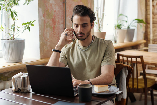 Młody mężczyzna freelancer w ubraniu casualwear rozmawia przez telefon komórkowy siedząc przed laptopem w kawiarni, siedząc w sieci i pijąc kawę - Zdjęcie, obraz