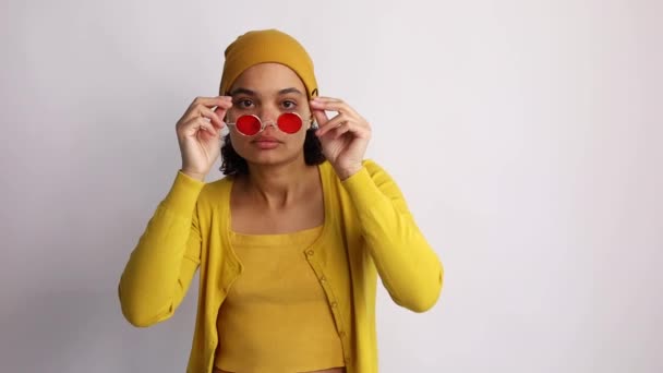 Retrato de chica africana fresca en gafas de sol con estilo rojo, suéter amarillo, mirando a la cámara en el fondo blanco de cámara lenta. Emociones de las personas.  - Metraje, vídeo