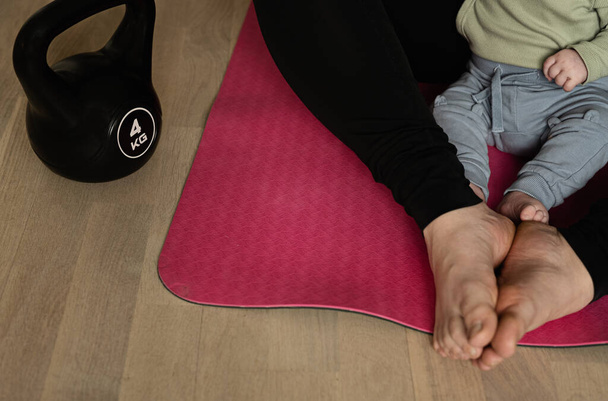 Baby und Frau sitzen auf Yogamatte bereit für Wochenbett-Training mit 4kg Kettlebell. Konzept des Trainings mit Baby oder Kleinkind. Aktiver Lebensstil mit Krafttraining nach der Geburt. - Foto, Bild