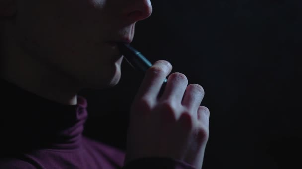 Sigara içmek için elektrikli cihaz. Karanlıkta bir adam elektronik sigara içiyor. Vape. E-sigara, vsping. Kavram: Sigara içmenin zararı. Yavaş çekim, karanlık arka planda sigara, yüzü olmayan bir adam elinde elektronik bir sigara tutuyor. - Video, Çekim