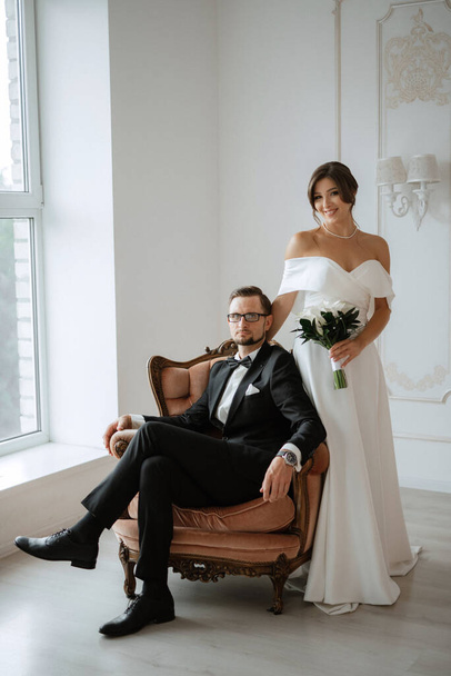 novio en un traje negro con corbata de lazo y la novia en un vestido blanco apretado en un estudio brillante - Foto, imagen