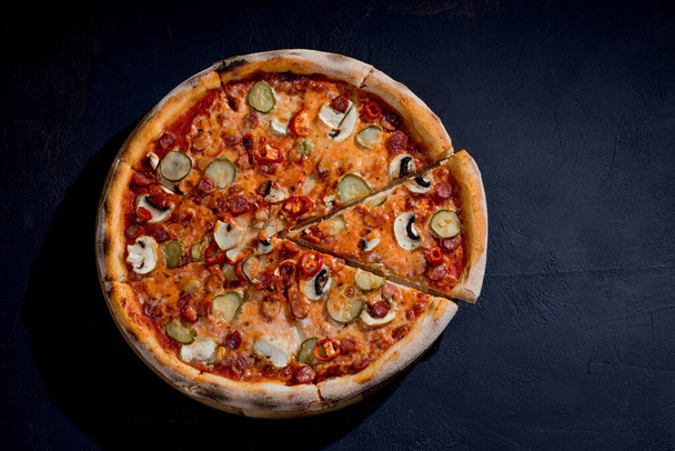 Appetizing pizza with hunting sausages, mozzarella cheese, pickled cucumbers, chili peppers on a tomato base on black background. - Photo, image