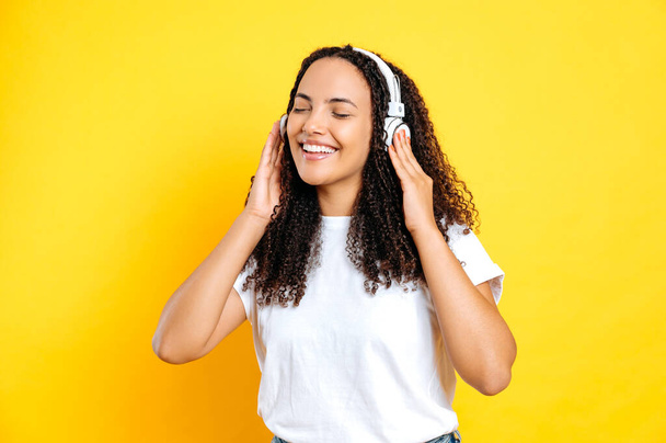 Cheerful pretty brunette brazilian woman, in casual stylish wear, with headphones, enjoy her favorite music, dancing with eyes closed, relaxing, having fun on isolated yellow background, smiling - Photo, image