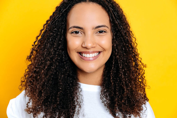 Detailní záběr fotografie šťastné nádherné pozitivní brazilské nebo hispánské mladé kudrnaté vlasy žena, na sobě bílé základní tričko, přátelský úsměv na kameru, stojí na izolovaném žlutém pozadí - Fotografie, Obrázek