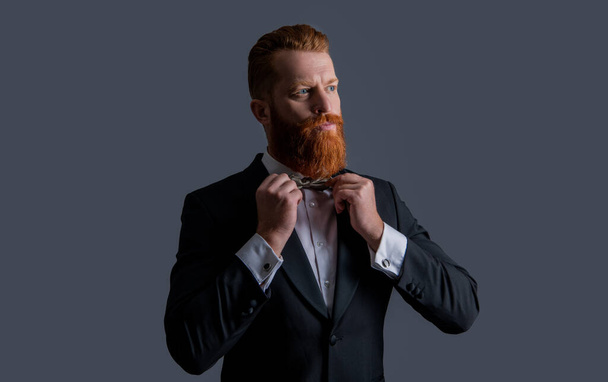 Elegant man fixing bowtie in black tuxedo. Handsome man with red beard studio. Bearded Irish man isolated on grey. - Photo, image