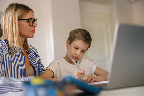Маленький целеустремленный школьник использует ноутбук, чтобы делать домашнее задание с помощью своей мамы  - Фото, изображение
