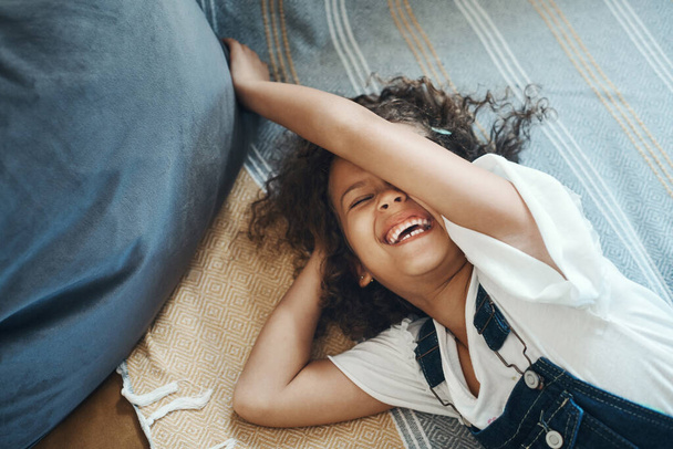 A house filled with laughing children is the best. an adorable young girl lying on the floor in the living room and laughing - Foto, afbeelding