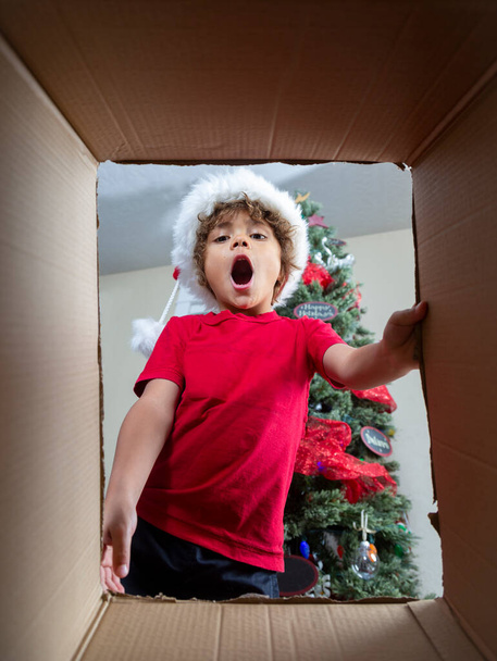 surpris garçon afro-américain préscolaire portant chapeau rouge de Noël et regardant à l'intérieur boîte en carton  - Photo, image