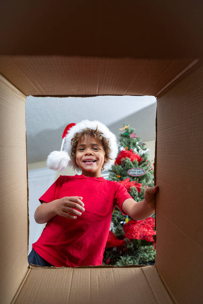 Happy diverse little boy opening a Christmas present on Christmas morning. View from below as he looks inside to see a gift he wants. Happy Holidays - Фото, изображение