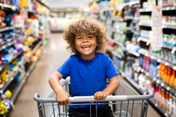 Lustiger kleiner Junge, der während eines Familienausflugs im Einkaufswagen sitzt und lacht. Er ist begeistert, etwas Neues zu kaufen - Foto, Bild