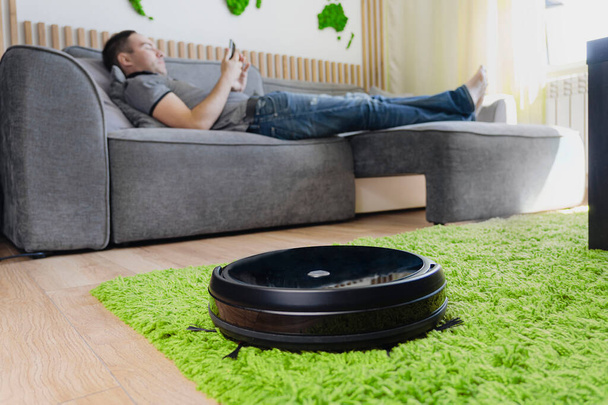Robot vacuum cleaner cleaning the living room. man enjoy rest, sitting on sofa at home. Remote control of robot vacuum cleaner via smartphone. Smart technologies at home. Internet of Things concept - 写真・画像