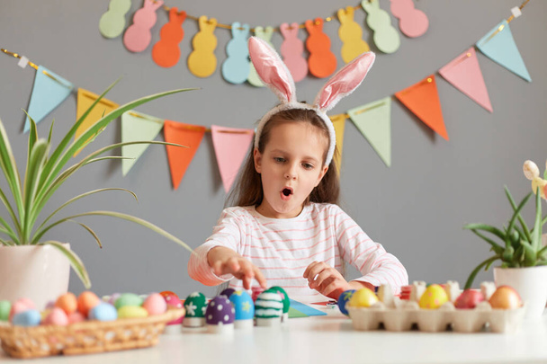 Image of amazed excited little girl dressed in rabbit ears sitting at table with Easter eggs, looking with open mouth and surprised face, preparing for celebration, posing against decorated gray wall - Fotó, kép