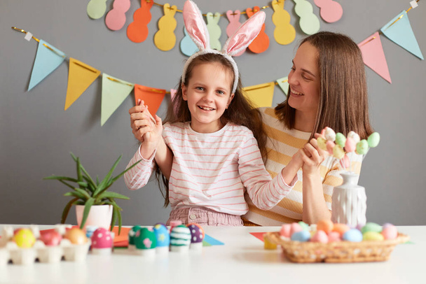 Horizontal shot of smiling little girl in bunny ears sitting with her mother at table among painted eggs, preparing for Easter, posing against gray decorated wall. - Foto, Imagen