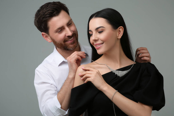 Man putting elegant necklace on beautiful woman against grey background - Фото, зображення