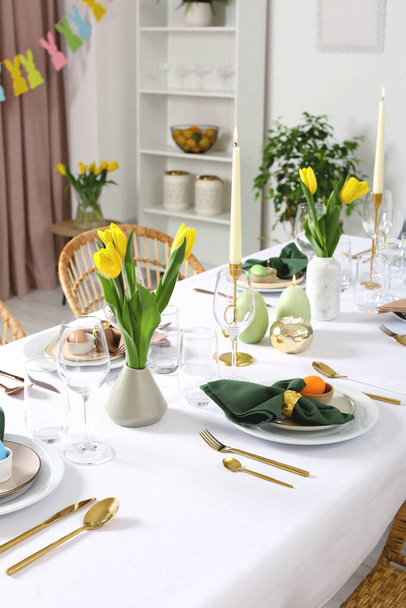 Festive Easter table setting with painted eggs, burning candles and yellow tulips in room - Foto, Imagem