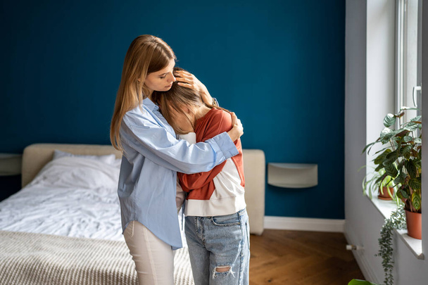 Loving caring mother hugging teen daughter. Parent mom showing understanding and support to upset sad teenage girl. Crying child sharing feelings with mommy while standing together at home - Fotografie, Obrázek