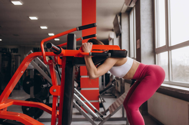 une fille avec une figure sportive squats dans la salle de gym sur un simulateur - Photo, image