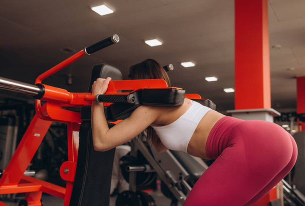 une fille avec une figure sportive squats dans la salle de gym sur un simulateur - Photo, image