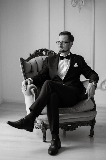 portrait of a groom in a black suit and glasses in a studio in an armchair - Foto, immagini