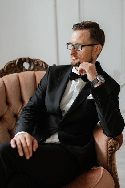 Porträt eines Bräutigams in schwarzem Anzug und Brille in einem Studio im Sessel - Foto, Bild